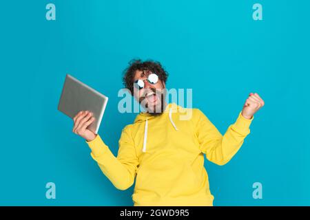 excited bearded man in yellow hoodie holding tab, screaming and celebrating victory on blue background in studio Stock Photo