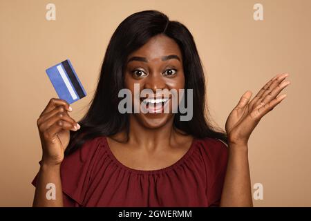 Joyful afro-american long-haired young lady holding credit card and cheerfully smiling at camera on beige studio background, going for shopping or cel Stock Photo