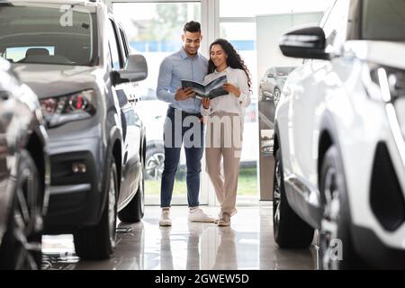 Cheerful arabic couple visiting luxury car salon together, handsome middle-eastern guy and beautiful long-haired lady reading brochure, checking auto Stock Photo