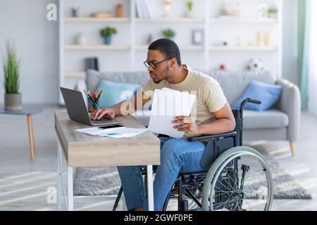 Paraplegic black guy in wheelchair with documents using laptop to work online from home. Handicapped young Afro freelancer with papers making business Stock Photo