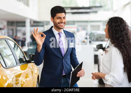 Attractive middle-eastern man luxury car showroom manager showing okay gesture and smiling while having conversation with long-haired brunette woman, Stock Photo