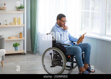 Stay home activities. Focused disabled African American guy with book sitting in wheelchair at home, empty space. Serious handicapped black man readin Stock Photo