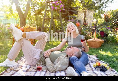 Romantic senior spouses enjoying picnic together, resting on blanket in garden and drinking red wine, chatting and laughing, enjoying weekend outdoors Stock Photo