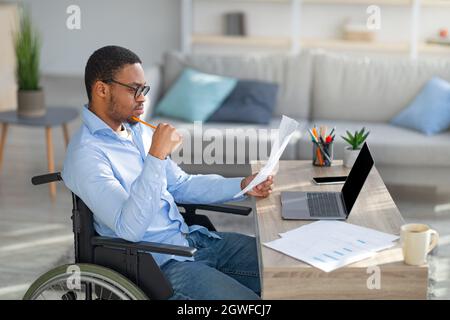 Focused black disabled man in wheelchair working with documents, using laptop at home office. Handicapped Afro man sitting at desk with computer, chec Stock Photo