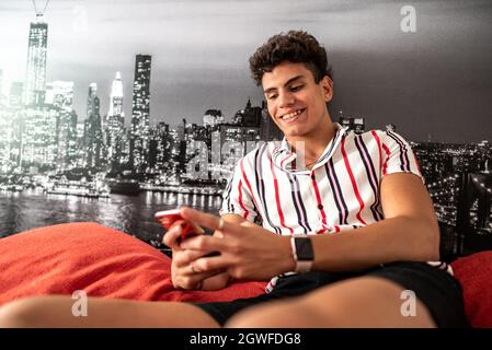 A close up shot on a happy Hispanic young guy smiling while using his phone in his room, Argentina Stock Photo