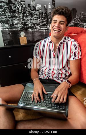 A close up shot on a happy Hispanic young guy laughing while using his laptop in his room, Argentina Stock Photo