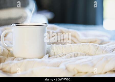 Hot steaming cup of coffee sitting on top a soft white knit blanket on a bed with stack of covers in background. Selective focus, blurred background. Stock Photo