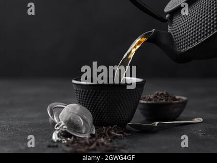 Pouring black tea from teapot to cup with strainer infuser and loose tea in ceramic bowl on black background. Stock Photo
