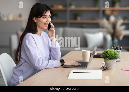 Portrait Of Positive Young Female Freelancer Talking On Cellphone And Working On Laptop Computer At Home Office, Having Pleasant Conversation, Enjoyin Stock Photo