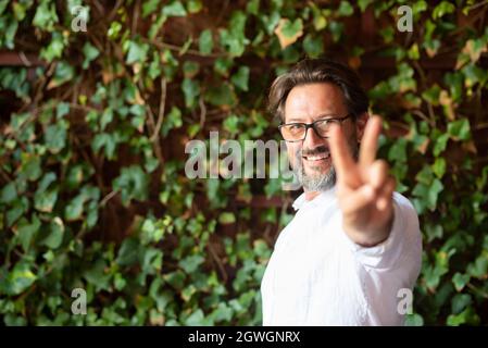 Happy mature man wearing eyeglasses and showing victory fingers sign while standing against creeper, Man showing v sign. Portrait of smiling man showi Stock Photo