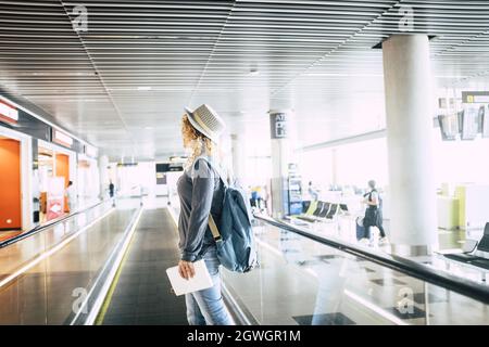 Young woman in hat going down by elevator at airport. Woman with backpack and holding digital tablet going for holiday trip. Profile of female passeng Stock Photo