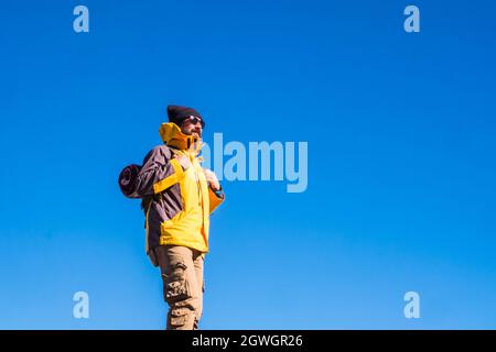 Mature man in backpack and sunglasses against blue sky. Male hiker in knit hat, sunglasses, backpack and yellow jacket admiring something interesting Stock Photo