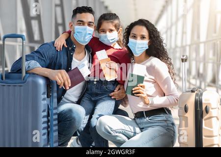 Family Travel During Pandemic. Arab Parents And Little Daughter Wearing Medical Masks In Airport Waiting For Flight At Terminal, Sitting Near Suitcase Stock Photo