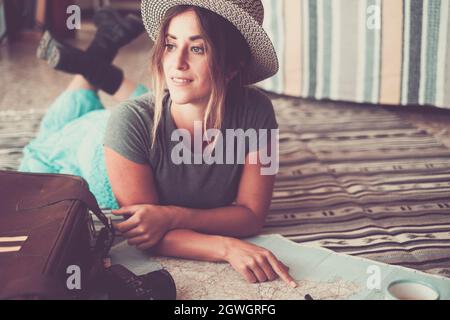 Beautiful young woman lying on carpet and pointing on atlas. Young woman in hat planning for location on map for vacation. Thoughtful woman daydreamin Stock Photo