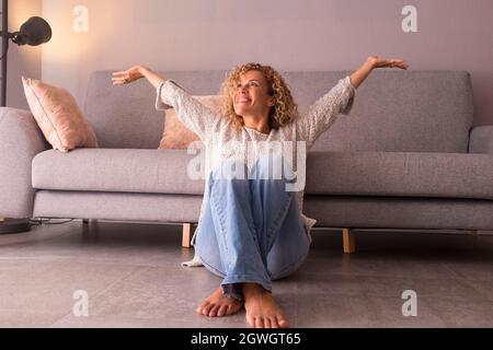 Cheerful young woman sitting on floor with arms raised at home. Thoughtful caucasian woman smiling sitting in living room. Satisfied woman sitting on Stock Photo