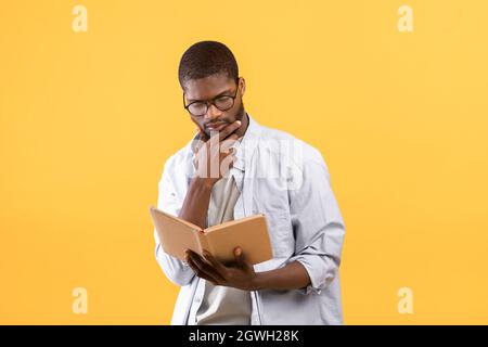 Thoughtful african american man in glasses reading book and touching chin, searching information in book for knowledge, standing over yellow backgroun Stock Photo