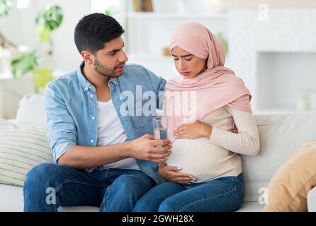 Care and support during pregnancy. Muslim expecting woman feeling sick, man giving her glass of water, siting on sofa at home interior, copy space. Ar Stock Photo