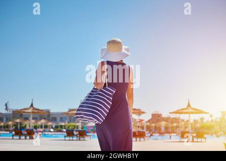 Woman In Hat And Blue Dress Go For A Walk In Hotel Resort Near Swimming Pool With Beach Bag