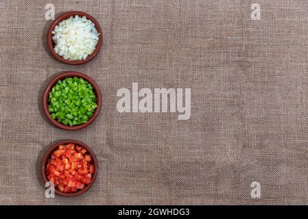 Background of rough fabric and brown color, with three small clay dishes with the ingredients of the sauce, onion, pepper and tomato.Copy space. Stock Photo