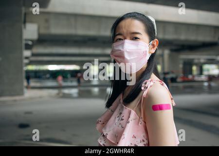 Portrait of smiling young Asian woman with covid-19 vaccination bandage standing wearing face mask for safety and precaution against coronavirus looking at camera Stock Photo
