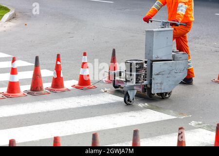 An orange overalls road worker renews pedestrian crosswalks on a road using a power trolley and traffic cones. Copy space. Stock Photo
