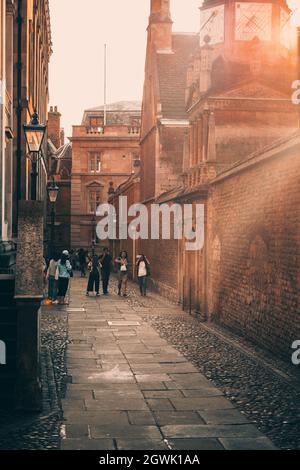 People walk along a pebble street among historical University Buildings decorated with lamp posts in Cambridge England Stock Photo