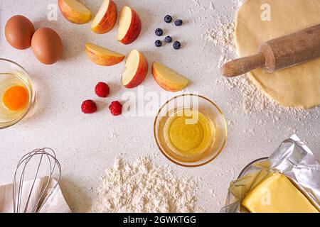 Table top view displaying raspberries, blueberries, apples, eggs, dough, butter and honey on a light kitchen counter surface Stock Photo