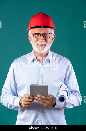 Portrait of senior man architect or construction engineer with helmet holding blueprints and tablet, against blue background Stock Photo