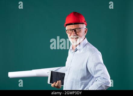 Portrait of senior man architect or construction engineer with helmet holding blueprints and tablet, against blue background Stock Photo