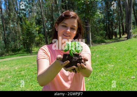Beautiful young Hispanic woman holding a small plant in her field hands before being planted in a green field surrounded by trees during the morning Stock Photo
