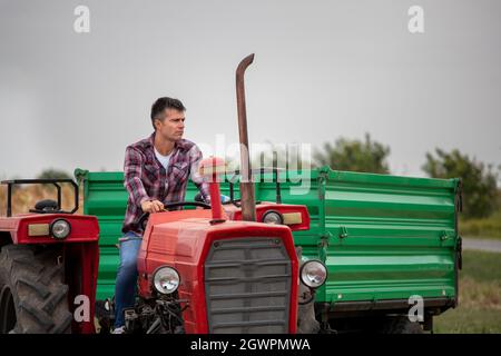 Attractive man smiling at camera in field in late summer. Handsome farmer driving tractor with trailer after harvest. Stock Photo