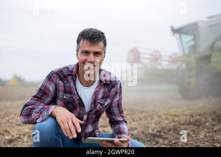 Young male agronomist using tablet in soy field. Smiling man crouching looking at camera with combine harvester in background. Stock Photo