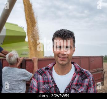 Attractive young agronomist standing in harvested field looking at camera. Two senior farmers filling tractor trailer with soy beans from harvester. Stock Photo