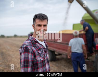 Attractive young agronomist standing in harvested field looking at camera. Two senior farmers filling tractor trailer with soy beans from harvester. Stock Photo