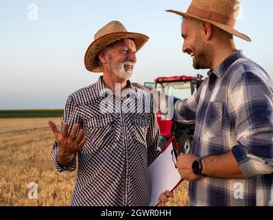 Elderly agronomist holding clipboard documents smiling. Young farmer holding shoulder laughing smiling. Two men standing in harvested wheat field with Stock Photo