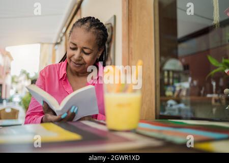 Senior african woman having fun reading a book during brunch time outdoor at bar restaurant - Focus on face Stock Photo