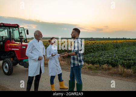 Elderly male and young female agronomists talking smiling using tablet wearing white lab coats. Young male farmer discussing agriculture with tractor Stock Photo