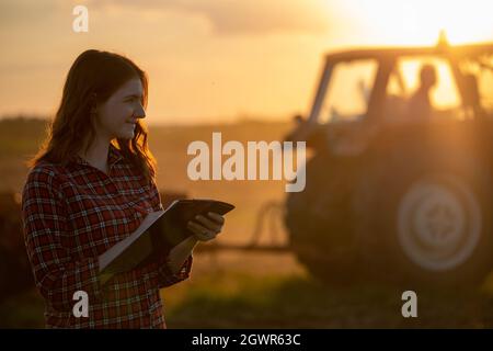 Portrait of attractive redhead farmer monitoring work in field at sunset. Agronomist taking notes on clipboard with tractor in background. Stock Photo