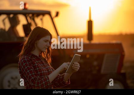 Portrait of pretty woman using tablet in agriculture. Young farmer standing in field in front of tractor at sunset. Stock Photo