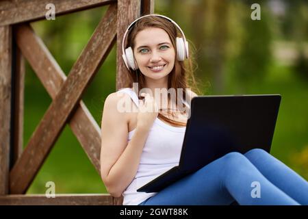 Woman in wireless headphones has telework with laptop outdoors. Stock Photo