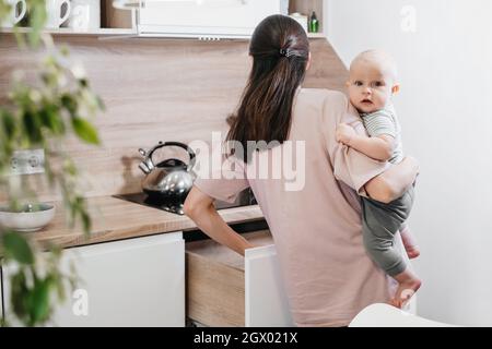 Busy mother cleaning kitchen with baby on her arms. Woman cooking and holding infant, tired woman doing daily homework with kid. Stock Photo