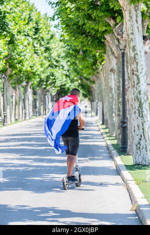 Cuban flag wrapped latino man riding an electric scooter along the street Stock Photo