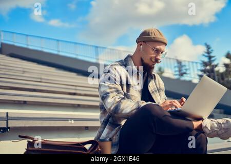 Serious bearded man in glasses works on pc, freelancer sits in park on stairs, manager on education Stock Photo