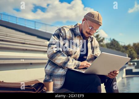 Serious student in glasses studies while sits near the university with laptop and headphones  Stock Photo