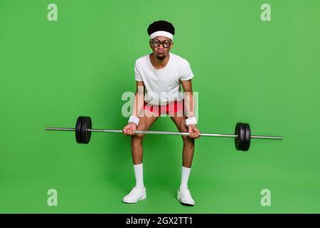 Photo of funny nerd bodybuilder guy lift heavy barbell grimace wear white t-shirt isolated green color background Stock Photo