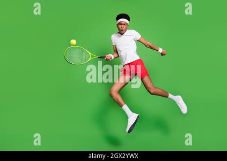 Full length photo of funky serious dark skin man wear white t-shirt jumping high playing squash isolated green color background Stock Photo