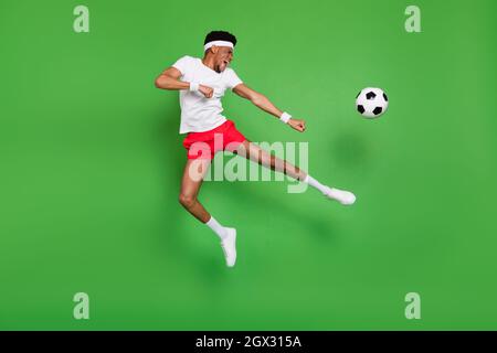 Full length photo of funky strong dark skin guy dressed sport outfit jumping playing football isolated green color background Stock Photo