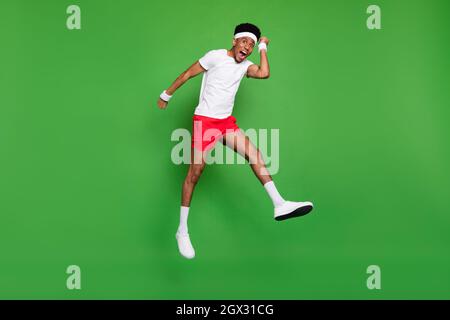 Full length photo of lucky funky dark skin guy dressed sport outfit jumping high rising fists isolated green color background Stock Photo