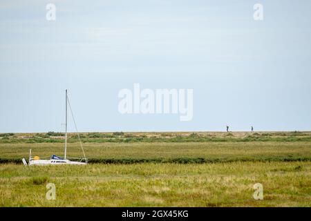 A couple walk on the bank of Blakeney beach with a dry river Glaven with a sailboat and marsh in the foreground. Norfolk, England. Stock Photo