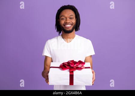 Photo of positive young afro american charming man hold hands gift box isolated on purple color background Stock Photo